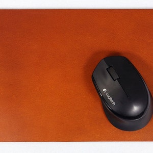 May include: A rectangular, brown leather mouse pad with rounded corners. A black Logitech wireless mouse rests on the pad. The mouse pad has a textured surface and is set against a white background.