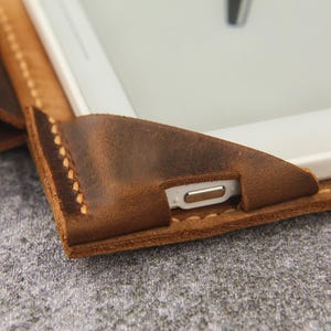 May include: Close-up of a brown leather tablet case with stitching detail. The case has a flap that covers the tablet screen and a cutout for the charging port.