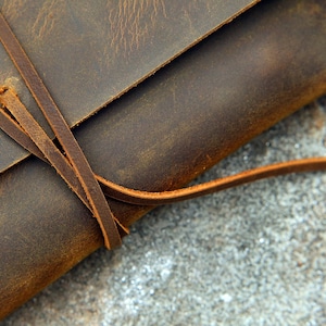 Op de afbeelding: Een bruine leren tas met een klep sluiting en een lange leren riem. De tas is dichtgebonden met de riem.