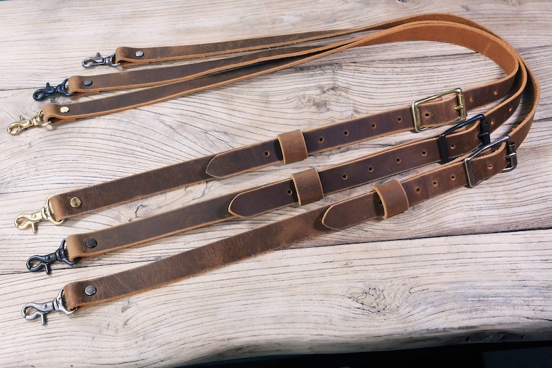 May include: Four brown leather camera straps with metal clasps and buckles. The straps are laid out on a wooden surface.