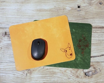 Pode incluir: Um mousepad laranja com uma ilustração de cachorro de desenho animado e um mouse sem fio preto. Um mousepad verde com um design decorativo é visível. Os mousepads estão em uma superfície de madeira.