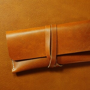 May include: A brown leather case with a strap closure. The case is made of a soft, supple leather and has a simple, elegant design.