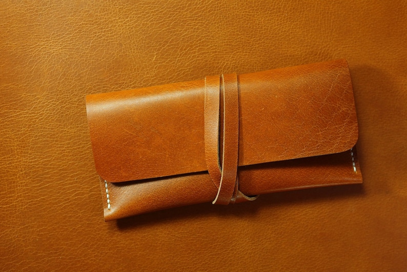 May include: A brown leather case with a strap closure. The case is made of a soft, supple leather and has a simple, elegant design.
