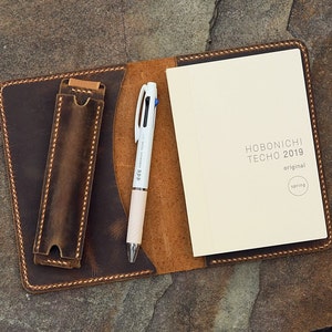 Peut inclure: Un journal en cuir marron ouvert avec un stylo et un carnet. Le journal a un porte-stylo. Un stylo blanc avec de l'encre bleue, noire et rouge est à côté d'un carnet crème. Le carnet indique "HOBONICHI TECHO 2019 original spring."