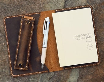 May include: Open leather journal with a pen and a notebook. The journal is brown with a pen holder. A white pen with blue, black, and red ink is next to a cream-colored notebook. The notebook reads "HOBONICHI TECHO 2019 original spring."