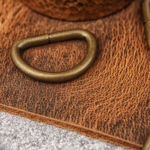 May include: Close-up of four antique gold-toned metal D-rings on a brown leather surface.