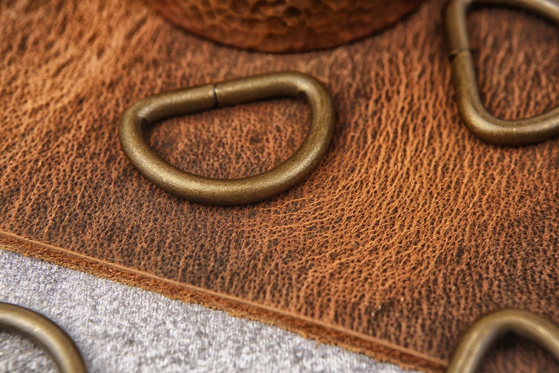 May include: Close-up of four antique gold-toned metal D-rings on a brown leather surface.
