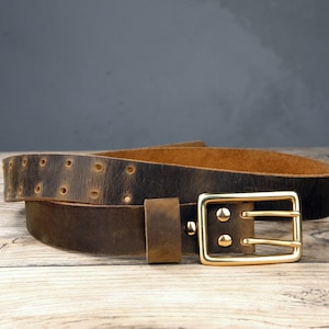 May include: A brown leather belt with a brass buckle. The belt has a distressed look and is folded in half. The buckle has a double prong design.