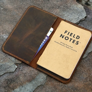 May include: A dark brown leather notebook cover with visible stitching along the edges. The cover holds a tan Field Notes memo book with black text that reads Field Notes, 48-Page Memo Book, Durable Materials / Made in the U.S.A. A card slot is visible.