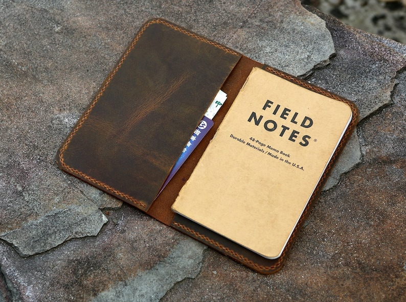 May include: A dark brown leather notebook cover with visible stitching along the edges. The cover holds a tan Field Notes memo book with black text that reads Field Notes, 48-Page Memo Book, Durable Materials / Made in the U.S.A. A card slot is visible.