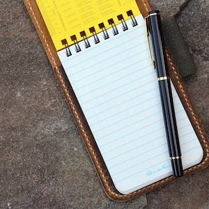 Op de afbeelding: Een spiraalgebonden notitieboek met wit gelinieerd papier en een gele referentiepagina met maateenheid-conversietabellen, in een bruine lederen hoes met zichtbare stiksels, vergezeld van een zwarte pen met goudkleurige accenten.