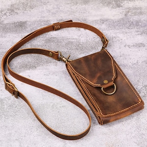 Könnte beinhalten: Braune Leder-Umhängetasche für Telefone mit einem langen, verstellbaren Riemen. Die Tasche hat einen Klappenverschluss mit einem Metallring und einem messingfarbenen Verschluss. Das Leder hat einen Used-Look, und die Nähte sind sichtbar.