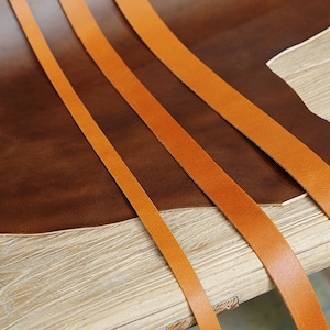 May include: Four strips of orange leather laid out on a brown leather surface. The leather is on a wooden table.