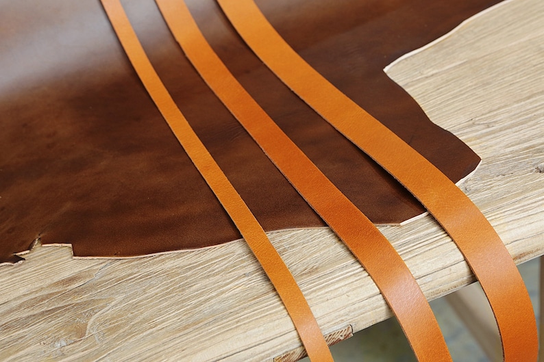 May include: Four strips of orange leather laid out on a brown leather surface. The leather is on a wooden table.