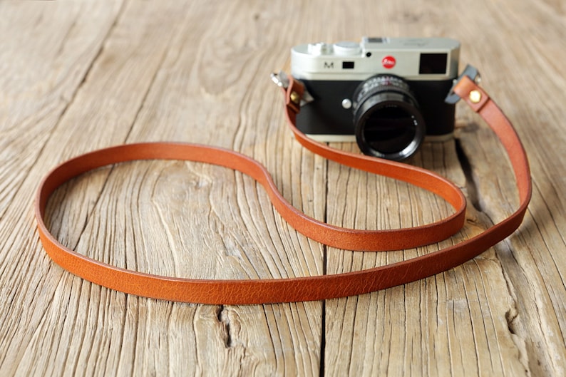 May include: A brown leather camera strap with a silver camera attached. The camera has a black lens and a red "M" on the top.