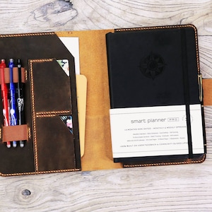 May include: A brown leather planner cover with a black planner inside. The planner has a compass design on the front and the text "smart planner PRO" on the back. The cover has a pen holder and a pocket for cards.