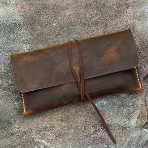 Op de afbeelding: Een bruine leren etui met een strik sluiting. De etui heeft een verweerde look en is gemaakt van dik, duurzaam leer.