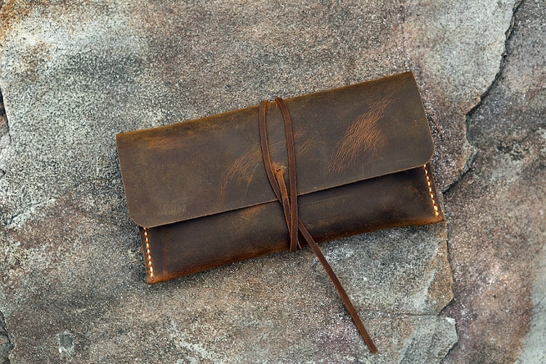 Op de afbeelding: Een bruine leren etui met een strik sluiting. De etui heeft een verweerde look en is gemaakt van dik, duurzaam leer.