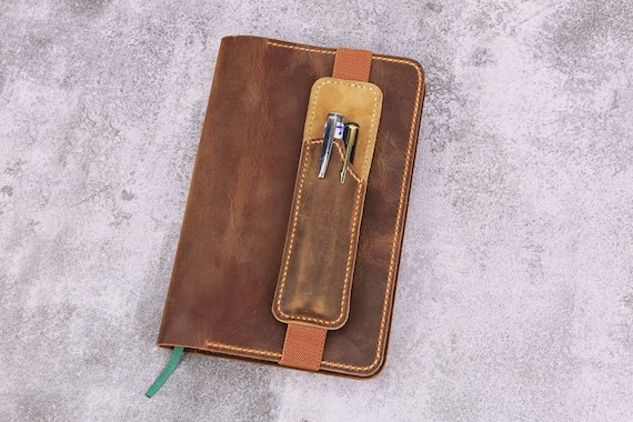 Hand-stitched leather wrap that clips directly onto a notebook or journal to keep your pen always at hand — a genuinely elegant analog solution.