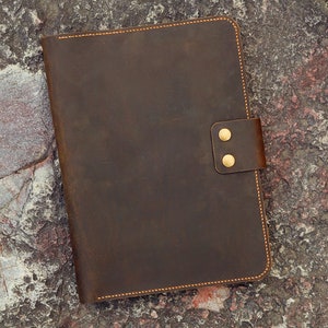 May include: A brown leather notebook with a flap closure and two brass poppers. The notebook is on a textured grey and brown surface.