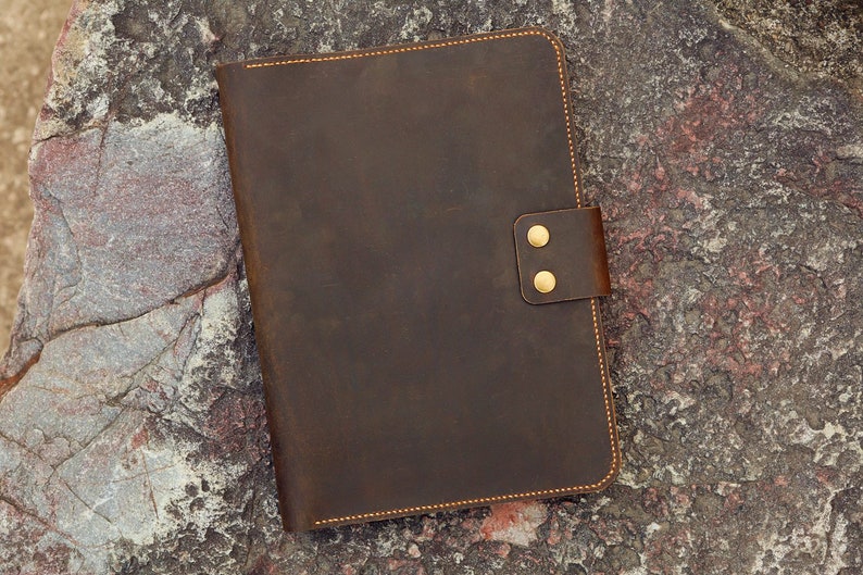 May include: A brown leather notebook with a flap closure and two brass poppers. The notebook is on a textured grey and brown surface.
