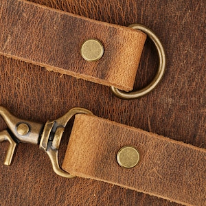 May include: Two brown leather straps with brass hardware, including a snap hook and a D-ring. The straps are on a brown textured surface.