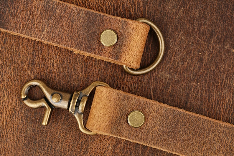 May include: Two brown leather straps with brass hardware, including a snap hook and a D-ring. The straps are on a brown textured surface.