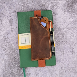 Puede incluir: Una cubierta de cuaderno de cuero marrón con un bolsillo para gafas y un portaplumas. La cubierta está unida a un cuaderno Moleskine verde con el texto "Moleskine" y "Cuaderno rayado" impreso en la parte delantera.