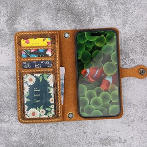 May include: A brown leather phone case with multiple card slots and a flap closure. The case has a floral pattern and a clear window to display the phone screen. The phone screen shows a picture of clownfish in a green and blue coral reef.
