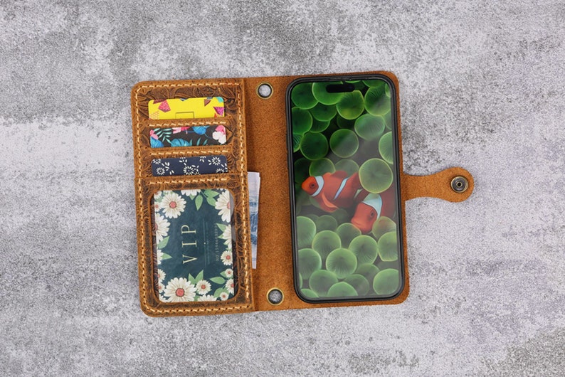 May include: A brown leather phone case with multiple card slots and a flap closure. The case has a floral pattern and a clear window to display the phone screen. The phone screen shows a picture of clownfish in a green and blue coral reef.