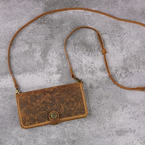 May include: A brown leather phone case with a floral pattern and a strap. The case has a flap with a button closure.
