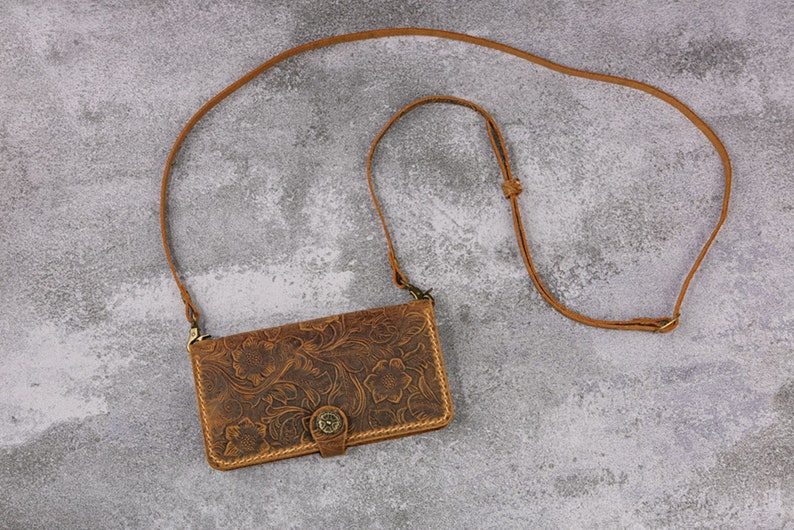 May include: A brown leather phone case with a floral pattern and a strap. The case has a flap with a button closure.