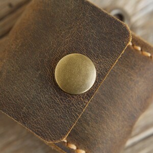 May include: Close-up of a small, brown leather pouch with a brass button closure. The leather has a distressed, textured appearance with visible stitching along the edges. The pouch is likely used for carrying small items.
