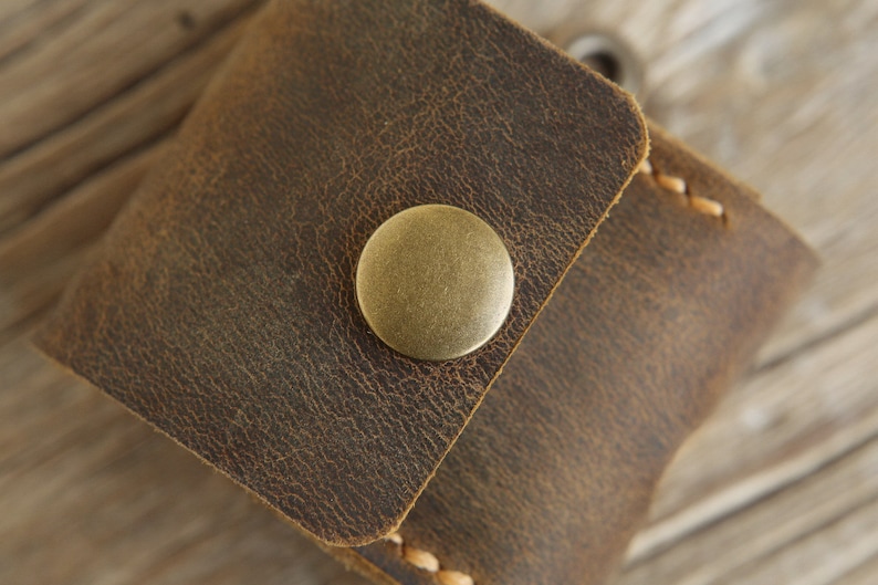 May include: Close-up of a small, brown leather pouch with a brass button closure. The leather has a distressed, textured appearance with visible stitching along the edges. The pouch is likely used for carrying small items.