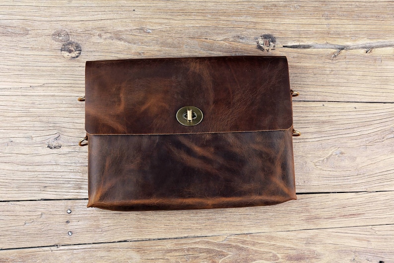 May include: A brown leather messenger bag with a brass clasp closure. The bag has a flap that folds over the front and is secured with the clasp. The bag is sitting on a wooden surface.