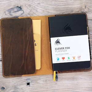 May include: A brown leather planner cover with a black planner inside. The planner has a fox logo and the text "Clever Fox Planner" and "Non-Dated Weekly Planner".