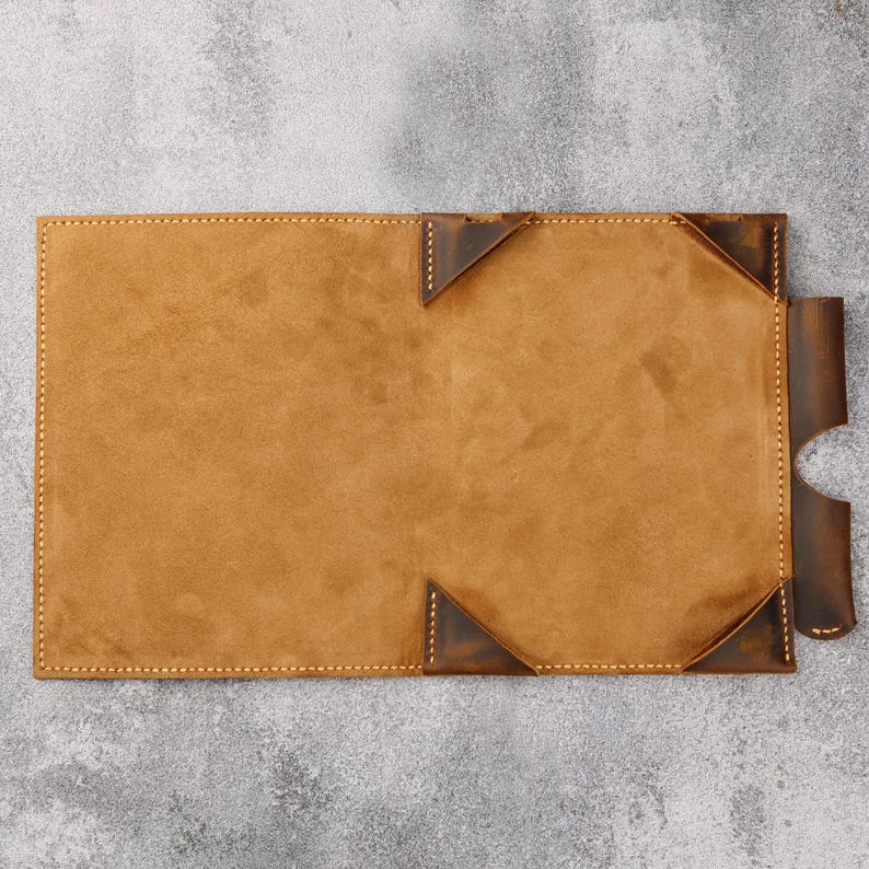 May include: A brown leather notebook with a stitched edge and a flap closure. The notebook is open and the inside is lined with a light brown suede material.