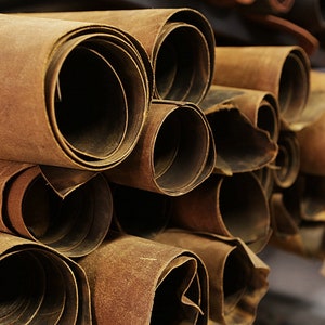May include: Close-up of a stack of rolled brown leather hides. The hides are tightly rolled, revealing the edges of the leather. The color is a warm, earthy brown, with variations in tone. The image suggests a craft supply or material for leatherworking projects.