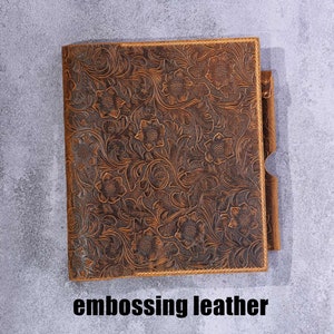May include: A rectangular brown leather journal cover featuring an intricate embossed floral pattern across the entire surface. The cover includes a side pen loop and the text embossing leather is printed in black below the object.