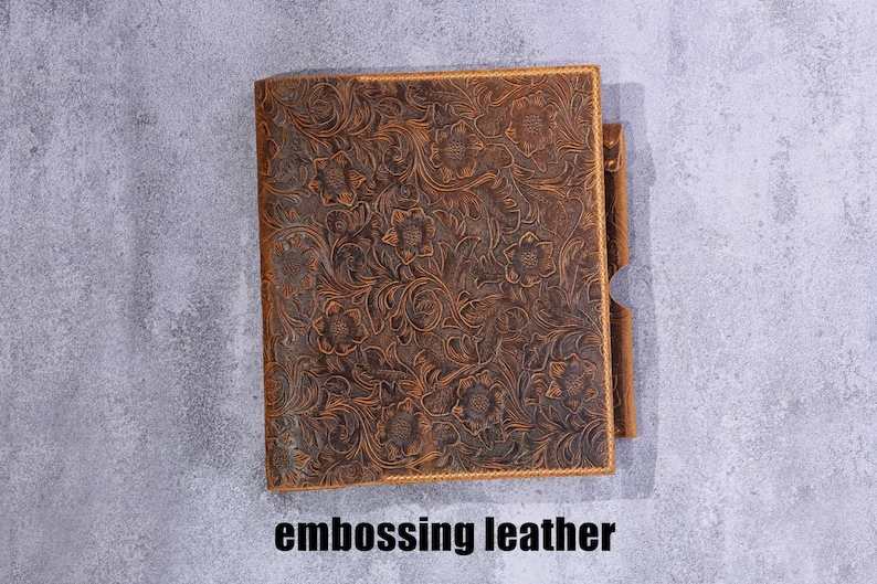 May include: A rectangular brown leather journal cover featuring an intricate embossed floral pattern across the entire surface. The cover includes a side pen loop and the text embossing leather is printed in black below the object.