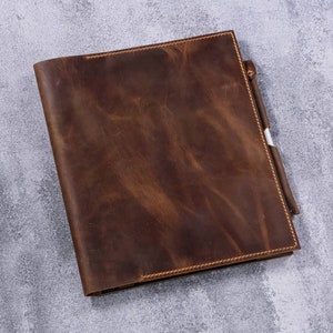 May include: A rectangular dark brown leather notebook cover with a smooth, distressed texture and visible tonal stitching along the edges. A matching leather pen loop is attached to the right side, holding a white pen with a silver clip.
