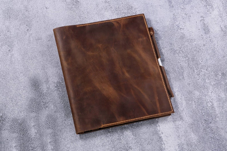 May include: A rectangular dark brown leather notebook cover with a smooth, distressed texture and visible tonal stitching along the edges. A matching leather pen loop is attached to the right side, holding a white pen with a silver clip.