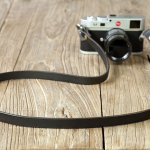 May include: A black leather camera strap with gold hardware, lying on a wooden surface. The strap is attached to a silver camera with a black lens.