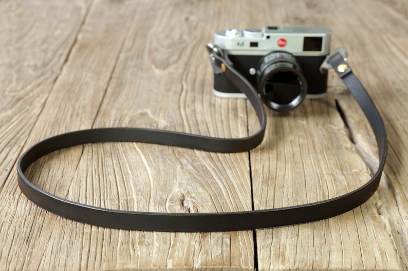 May include: A black leather camera strap with gold hardware, lying on a wooden surface. The strap is attached to a silver camera with a black lens.