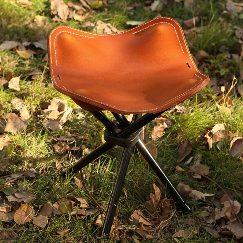 Puede incluir: Un taburete de camping de cuero marr&oacute;n con un marco de metal negro en forma de tr&iacute;pode. El taburete est&aacute; sentado en una cama de hierba verde y hojas marrones.