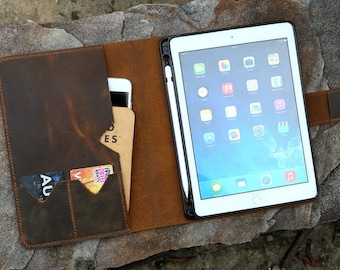 May include: A brown leather iPad case with a built-in stand and card slots. The case holds a white iPad with a black border and a stylus. A smartphone and a notepad are also visible inside the case.