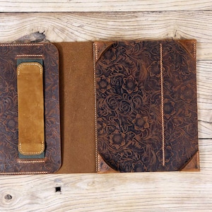 May include: A brown leather folio case with a floral embossed design. The case has a flap that closes with a snap and a handle on the side. The case is open to reveal a brown leather interior. A matching brown leather strap is included.