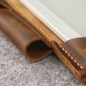 May include: Close-up of a brown leather tablet sleeve with stitching detail. The sleeve is folded over to show the interior lining.