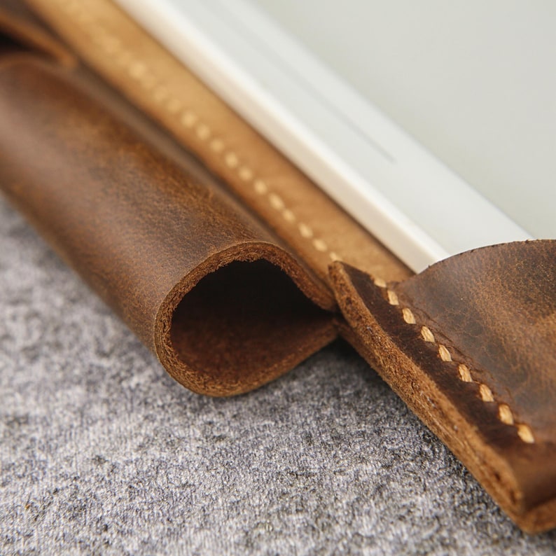 May include: Close-up of a brown leather tablet sleeve with stitching detail. The sleeve is folded over to show the interior lining.
