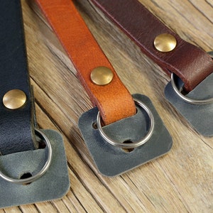 May include: Three leather camera straps with brass accents and metal rings. The straps are black, brown, and tan.